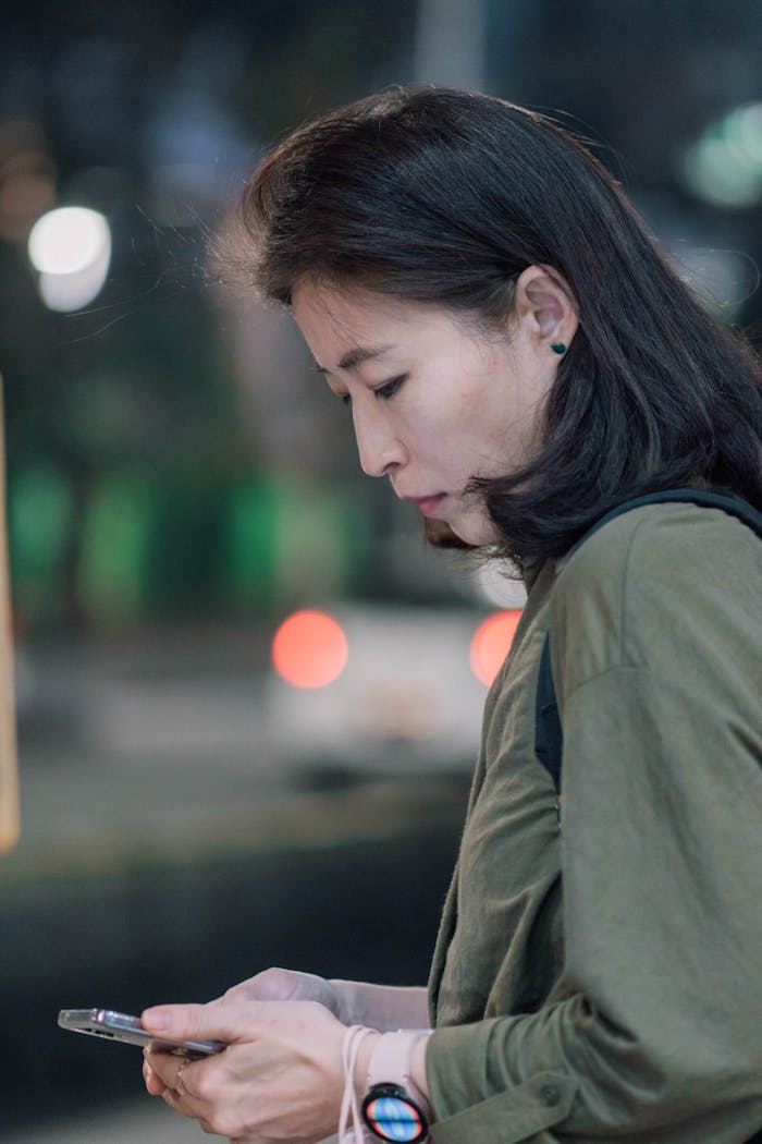 A woman with black hair is looking down while using a smartphone outdoors at night.
