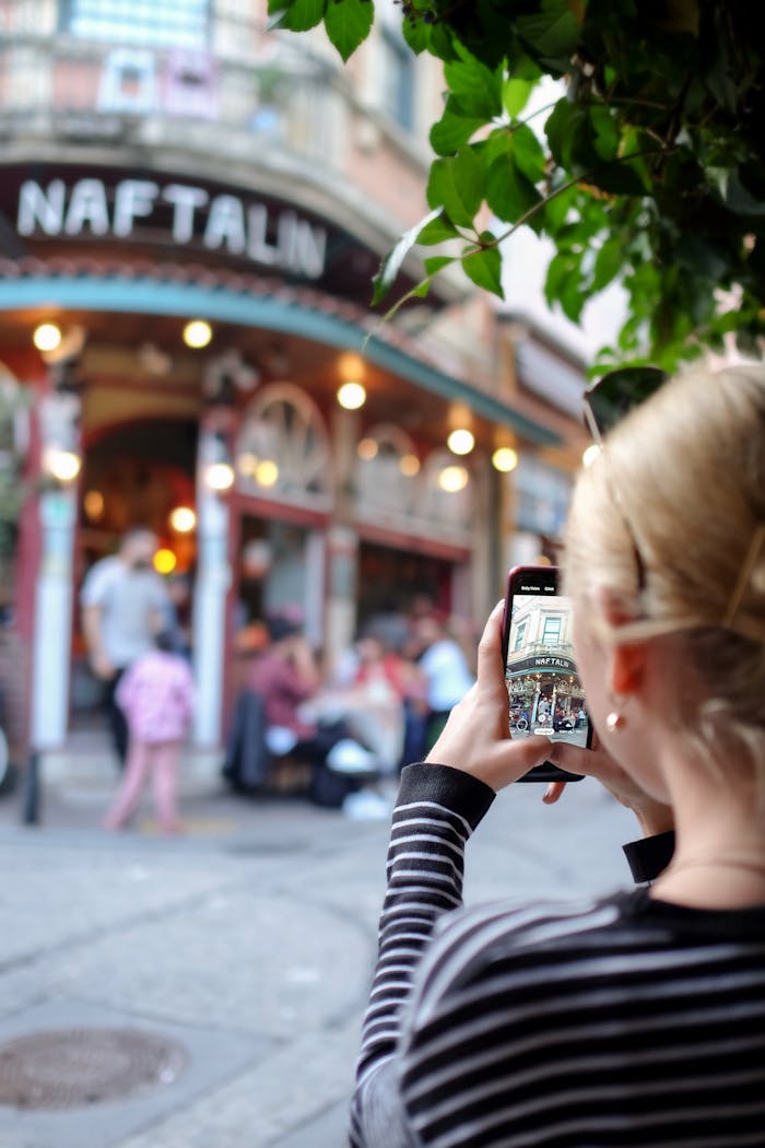 Back view of crop unrecognizable female photographer taking picture of building on smartphone located on street with people in city